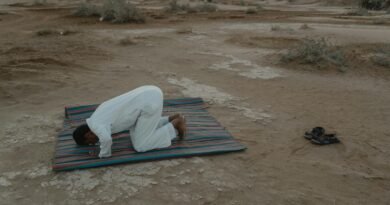 A Muslim man kneels in prayer on a colorful mat in the desert during sunset, showcasing a serene moment of faith.